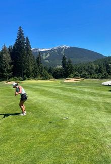 Michela Cava swinging a golf club on a lush green golf course with mountains in the background.