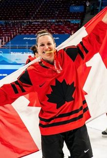 Jill Saulnier smiling with a gold medal in her mouth and holding a Canadian flag behind her back