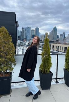 Abby Newhook in a black coat smiling on a rooftop with a city skyline in the background.