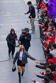 Mannon McMahon in a black skirt and boots, high-fiving fans behind a barrier. A camera crew follows her.