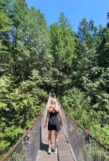 Mannon McMahon in a black dress walks across a narrow suspension bridge surrounded by lush green trees under a clear blue sky.