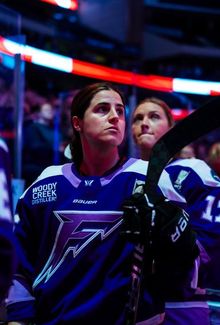 Michela Cava standing on the bench with her teammates with her helmet off for the national anthem.